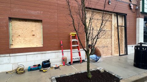 Storefront windows boarded with plywood by Stonewing after glass damage, ladder and tools on the sidewalk